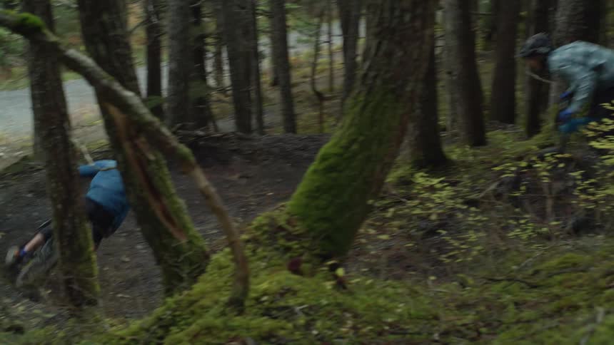 Two friends ride mountain bikes around a curve in a dirt trail through the trees