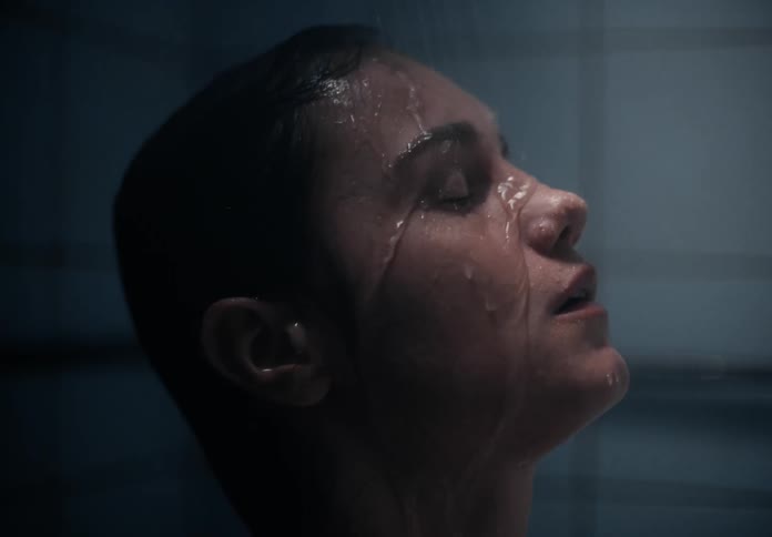A young woman stands in the shower with her eyes closed as water runs over her face