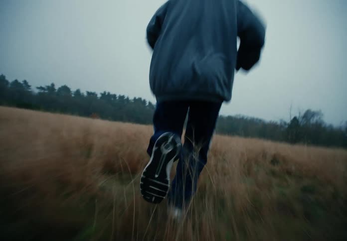 A low-angle handheld shot following a young woman jogging through a grass field during athletic training