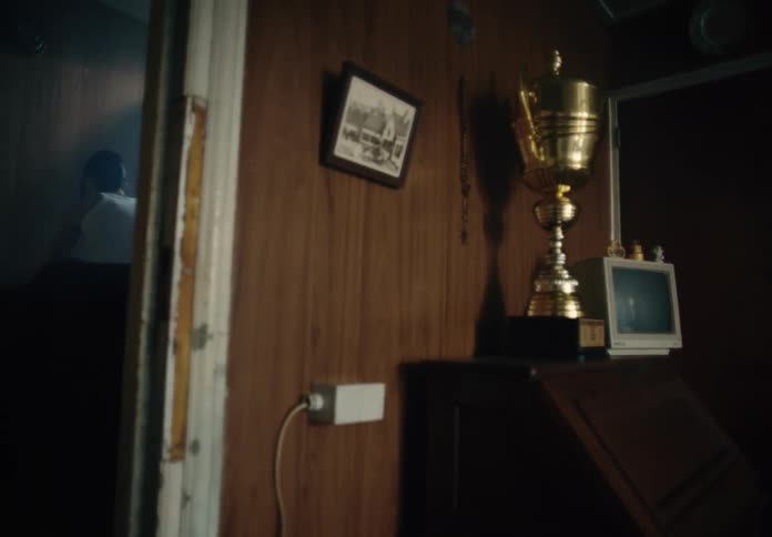 A large trophy sits on a writing desk outside a bedroom where a young woman sits leaning forward