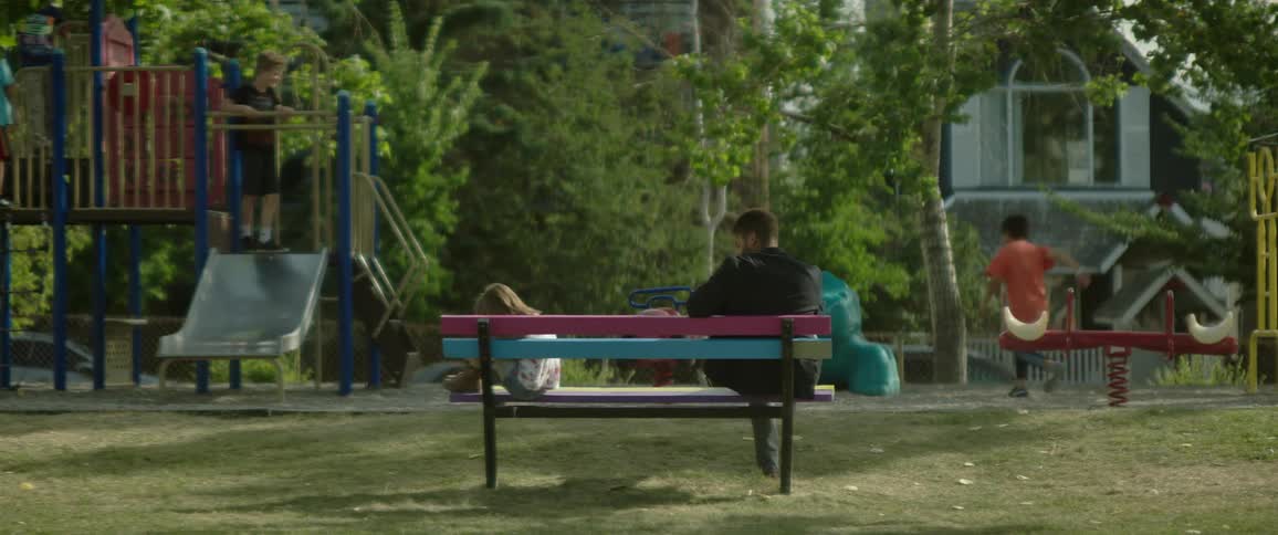 A little girl is sitting alone on a park bench holding her knees when a man walks by and sits down near her as other children are playing