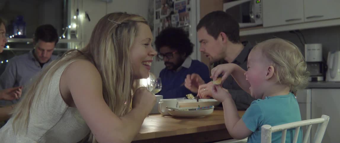 A woman seated at a dinner table with several other people laughs and talks with a toddler