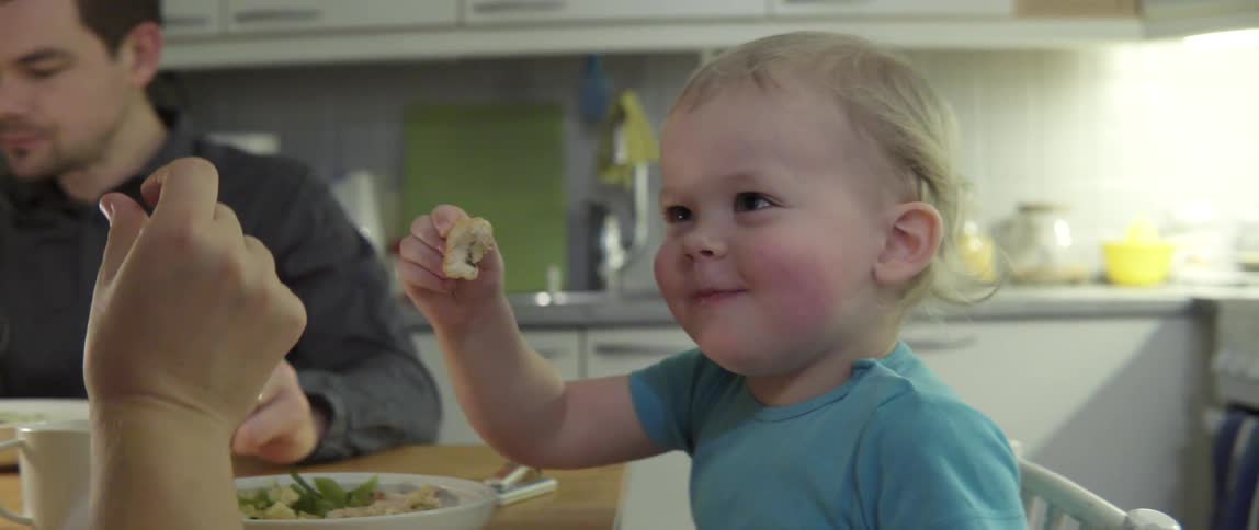 A baby laughs and smiles as he eats food at a dinner table