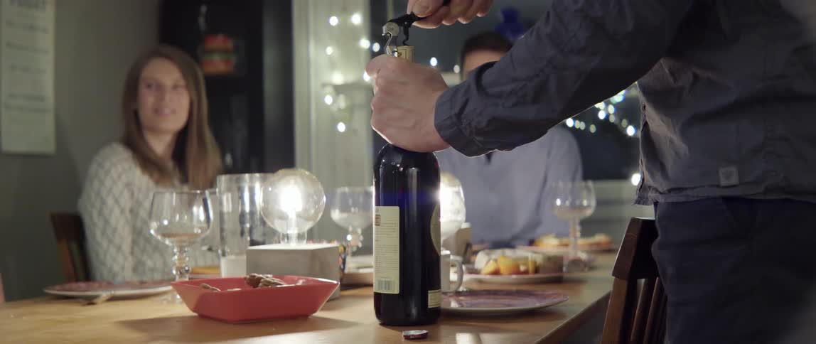 Young man preparing to serve wine to friends seated around a modern indoor dinner table
