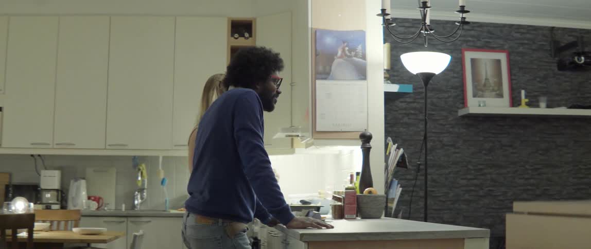 A man and woman stand in a kitchen at night and speak to others off camera