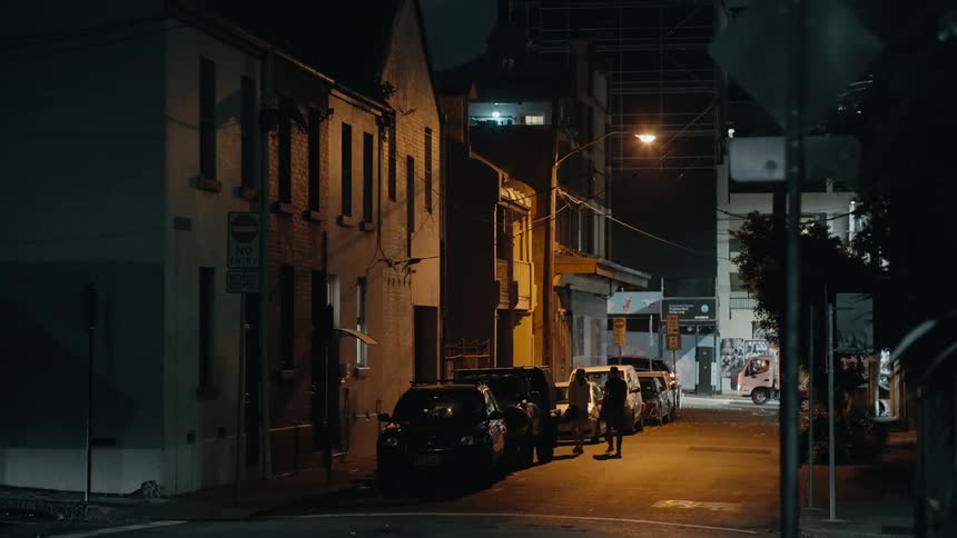 A man and woman cross a dimly lit street together in a city at night
