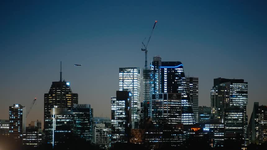 An airplane flies over lit up high rise office buildings and construction cranes at night