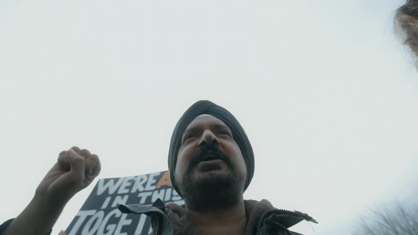 Man raises hands in air at protest rally as someone waves sign in background