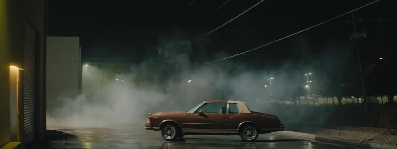 Smoke flows across parking lot near where sedan is parked at night