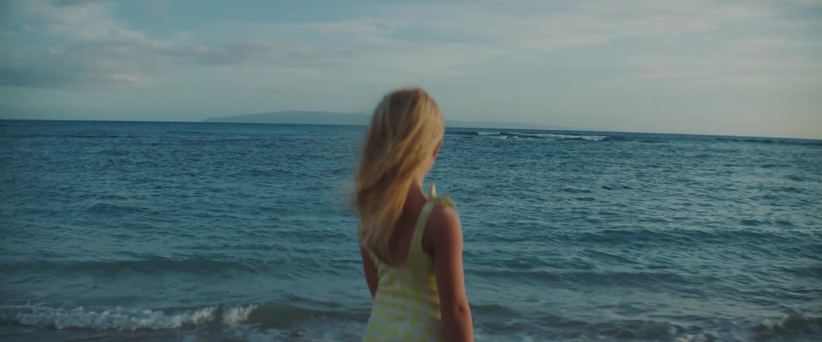 A little girl is standing on the beach watching the waves roll in as the wind blows her long blond hair