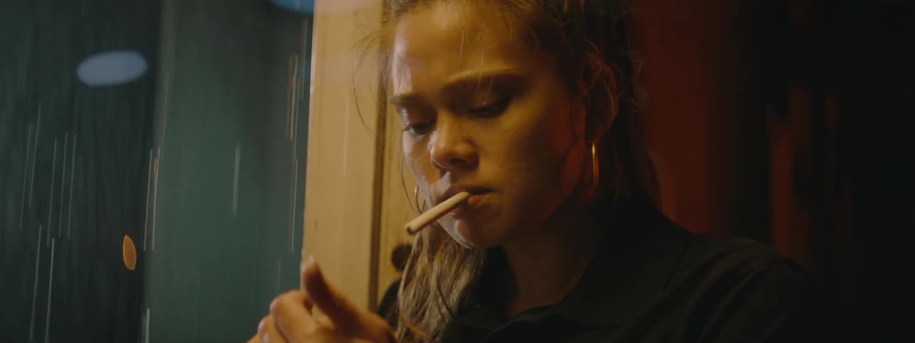 A woman stands by a door smoking a cigarette with it raining outside