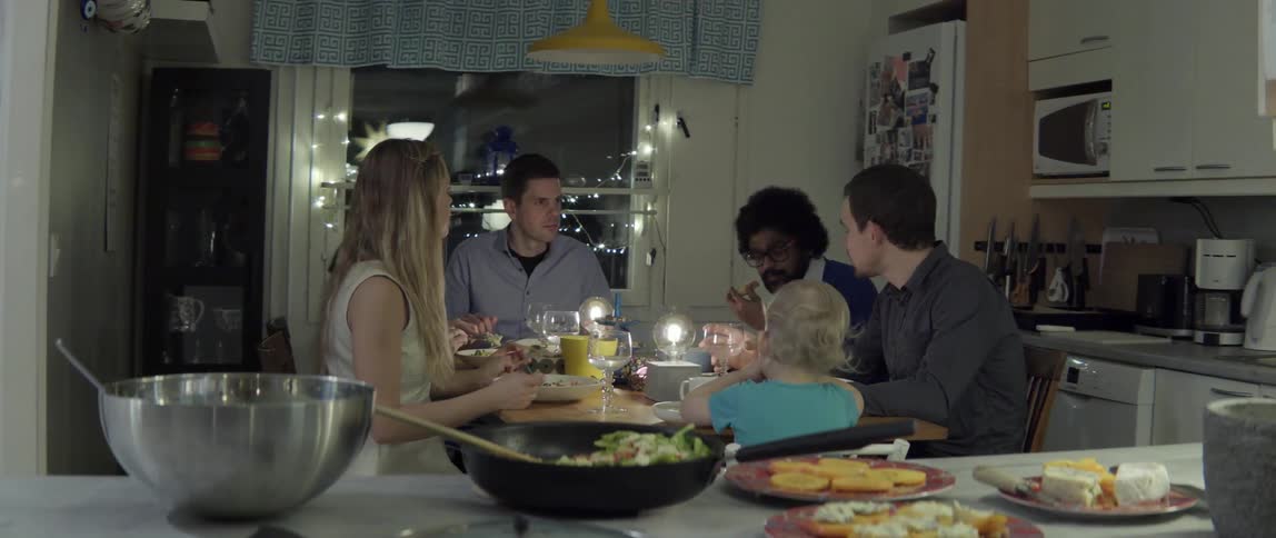 A group of adults and a toddler sit at a dinner table in an apartment at night