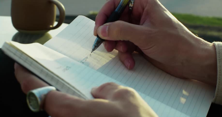 A mans hand is seen holding a pen and writing in a book and there is a coffee cup on a table nearby