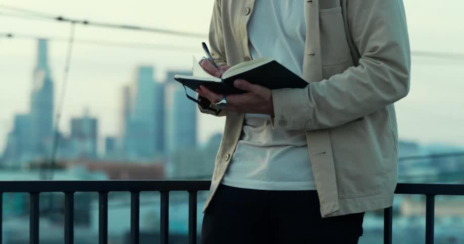 Man leans against rail while writing in journal with city skyline in the background