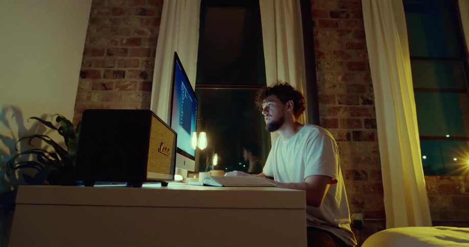 A man sits at a computer desk in his home looking at a large monitor