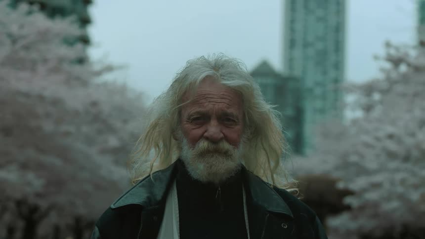 An elderly man looks into the camera while standing outdoors during the day