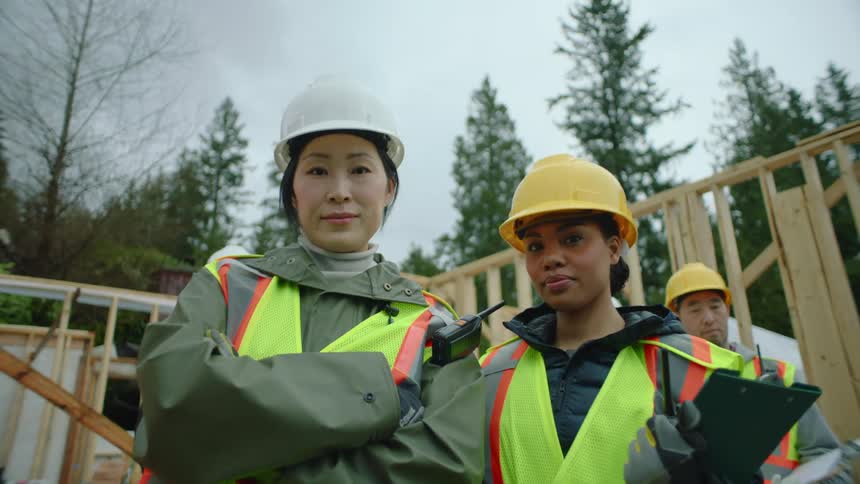 Two female construction workers stand in front of a building frame