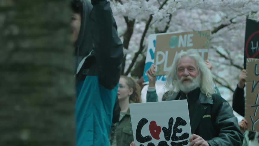 Protesters holding signs march under flowering trees near buildings with landscaping