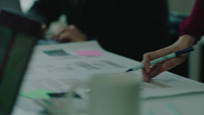 Woman stands at meeting explaining things on sheet as another employee takes notes on a laptop