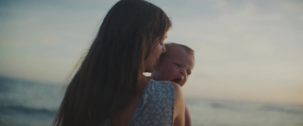 Mother holding and taking care of her baby in front of the sea at the beach during sunset