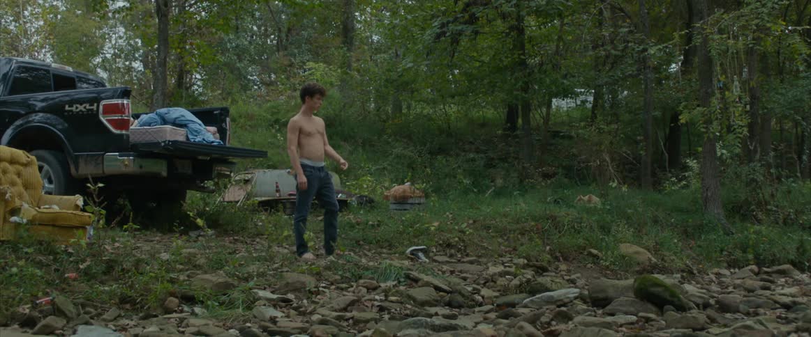 A teenage boy sits in the back of a truck parked in a forest with a mattress in the back of the truck and gets out and walks and then a young woman sits up and follows him