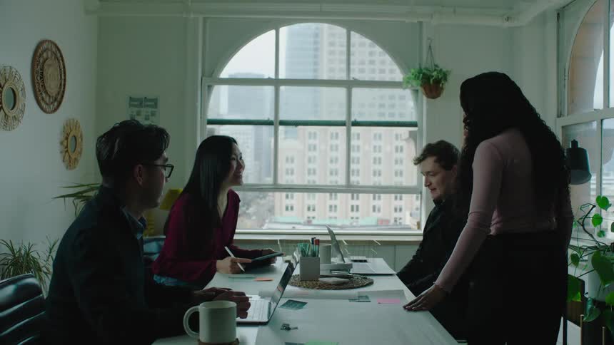 A woman walks in to an office where coworkers are on laptops and tablets and talks with them and looks at what they are working on
