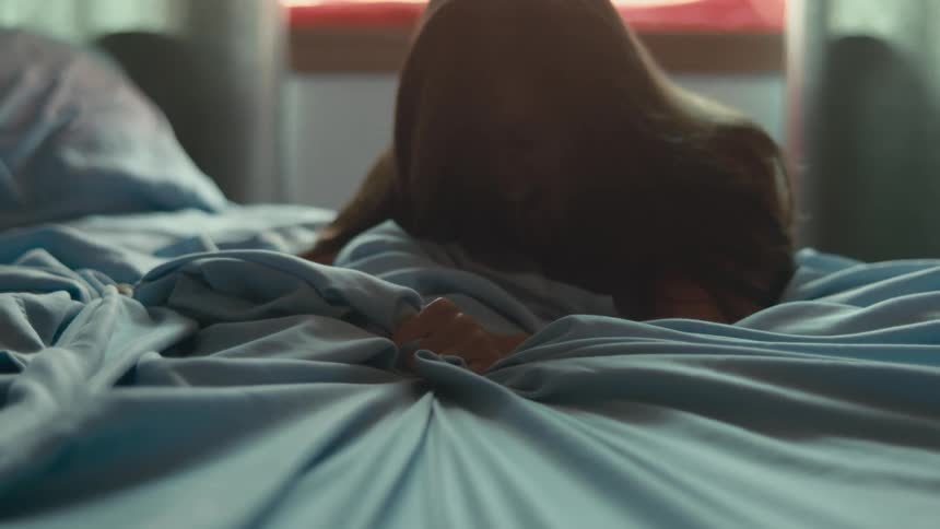 A woman kneeling beside a bed grimaces and yells as she grabs and pulls sheets and hits the bed