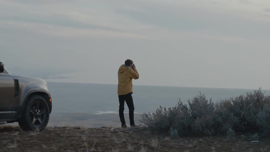 A man gets out of a vehicle parked on a cliff and takes photos of the land and water in front of him