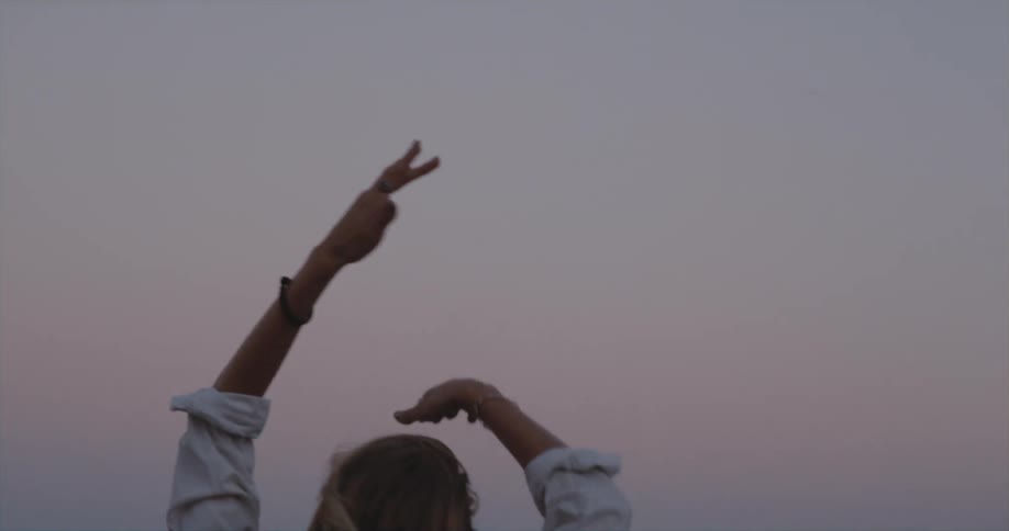 Blonde woman with black vest and white shirt dances on the beach at dusk