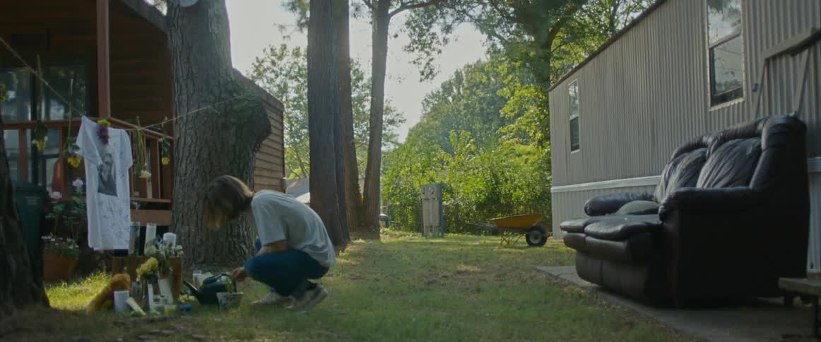 A young man waters a memorial plant and then sits in a couch outside a home