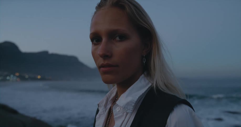 Woman stands on shore as waves roll in and town lights twinkle in the distance