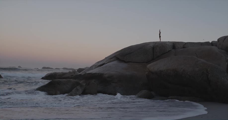 Silhouette of a person standing on shore along the coast as waves splash and crash into rocks below