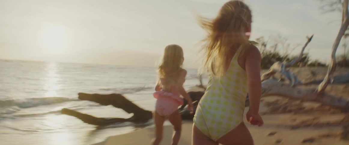 Three young children chase each other around tree branches on a quiet beach