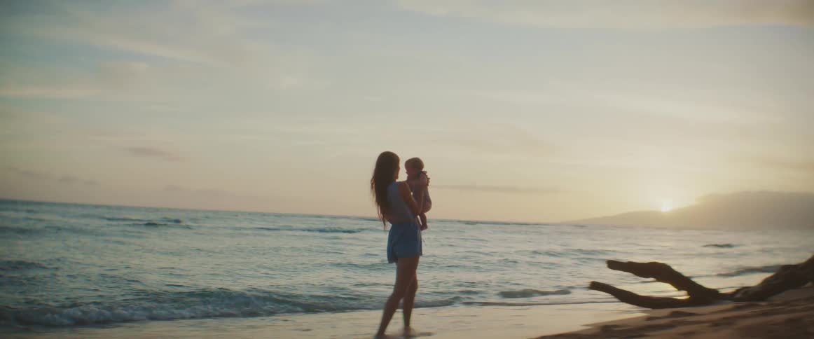 A woman is on the beach and she tosses her baby into the air as the waves roll in