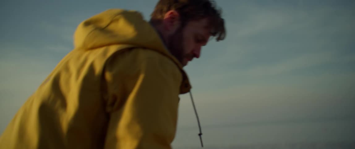 A man kneels on a beach to pick up a stone and throw it out across the water