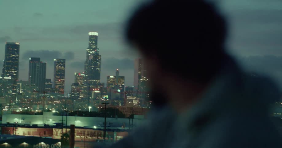 Man with beard and jacket looks at illuminated skyscrapers at dusk