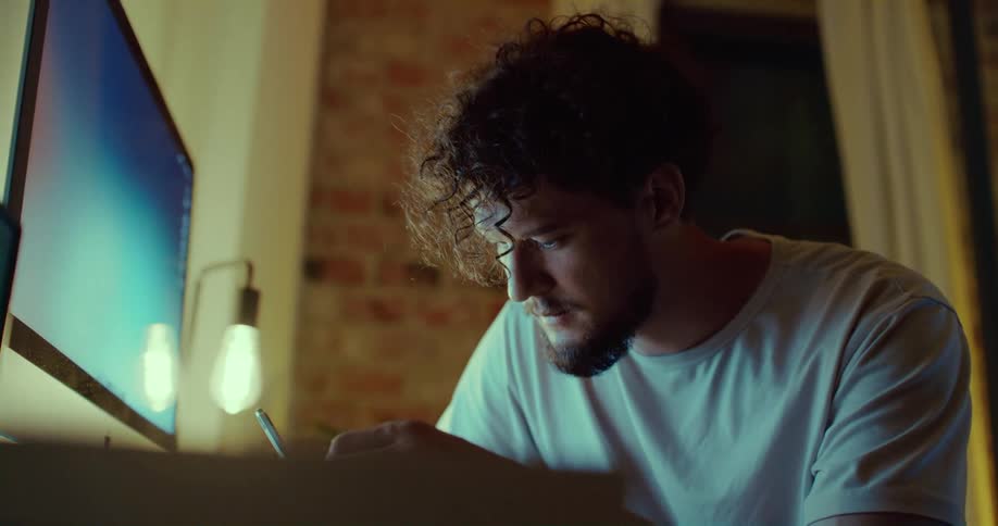 A man sits in front of his computer at home and writes in a book