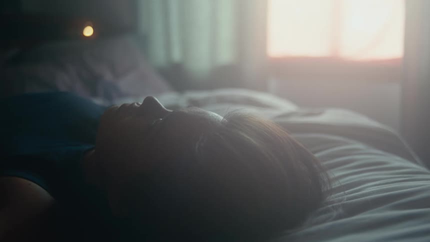 A woman lies across a bed looking out the window and then to the ceiling as she brings her fist to her forehead upset