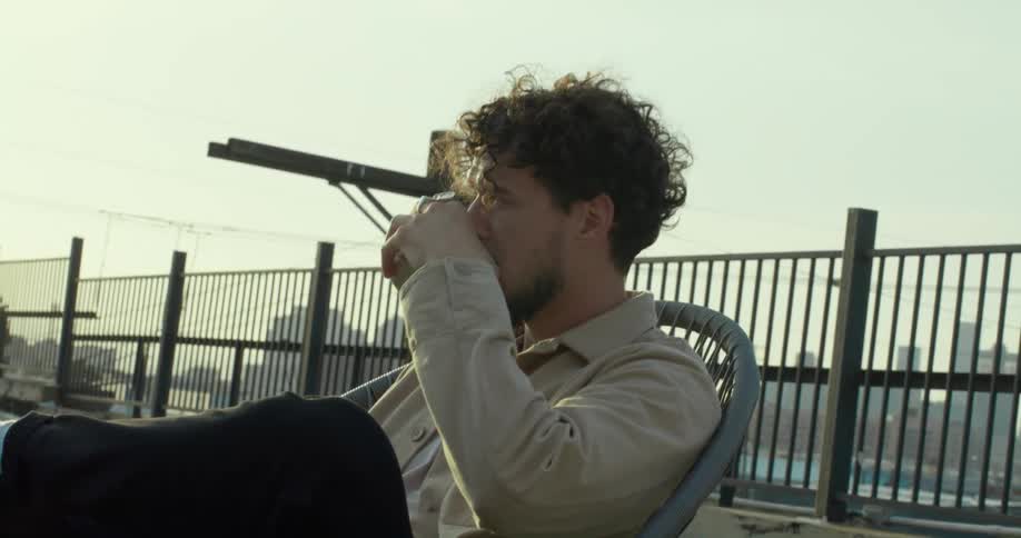 A man sits on a rooftop terrace and takes a drink from a cup
