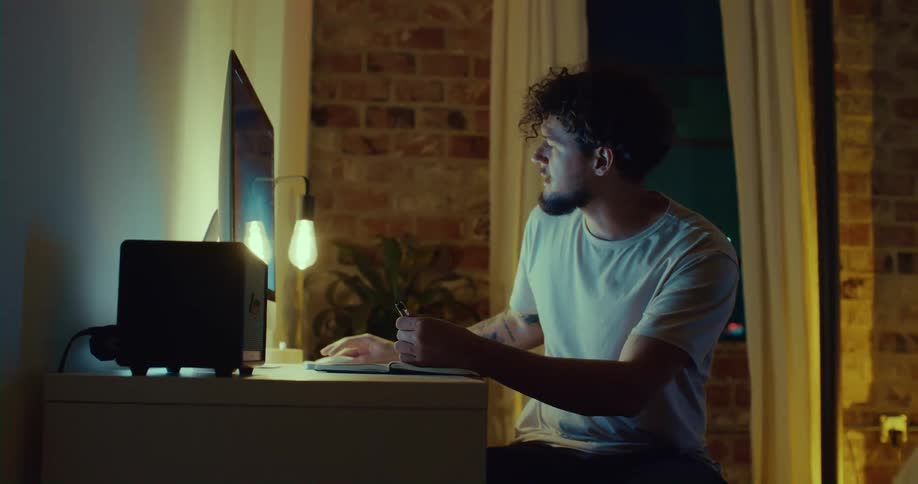 A man is sitting at a desk looking at a computer monitor and writing something in a book