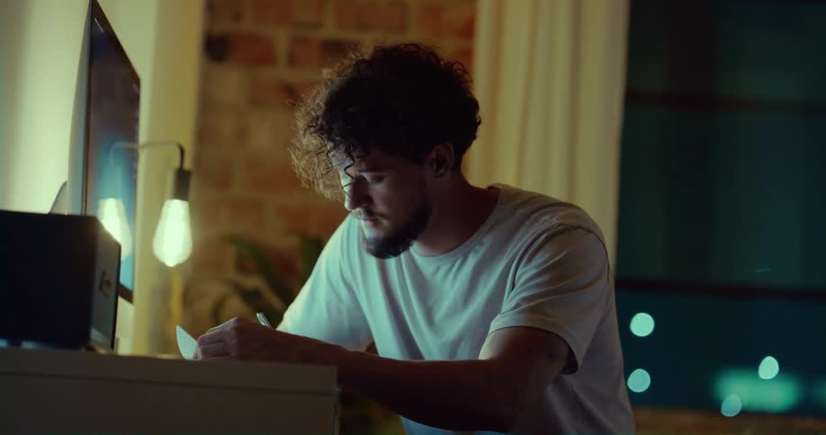 A man is sitting at a desk looking at a computer monitor and writing in a book