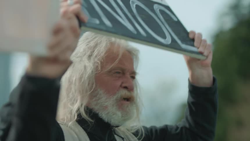 A man holds up a sign and chants while protesting outdoors in a city on a cloudy day