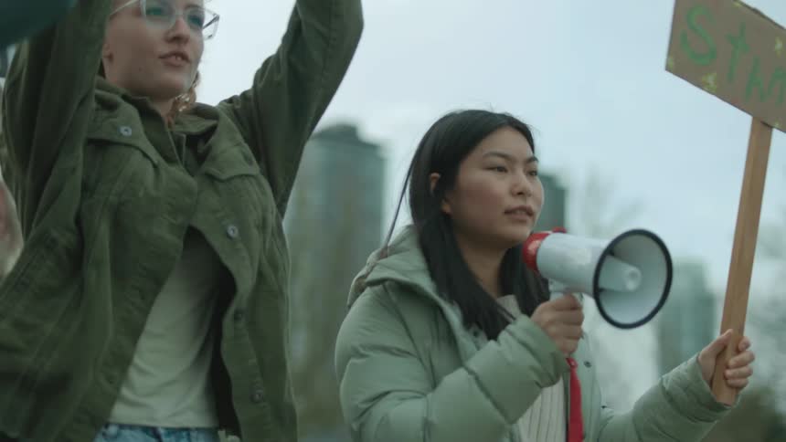 A group of demonstrators are holding signs and an asian woman is holding a megaphone as they march down the street