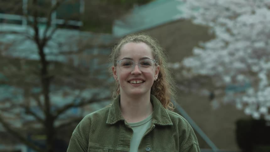 A caucasian teenage girl stands smiling and laughing in front of a school building