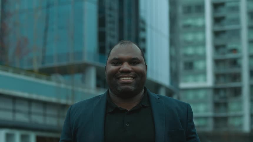 Black man wearing a suit standing outdoors in front of high buildings and smiling
