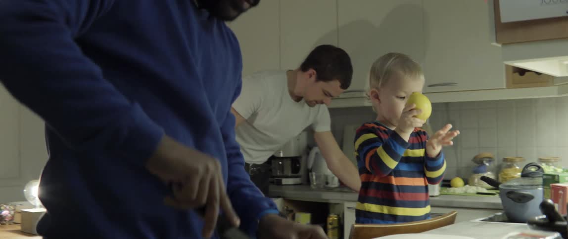 Two men work in the kitchen while a toddler stands on a chair at the counter