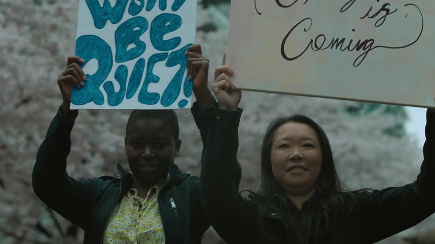 Two female protestors hold signs up while standing near flowering trees with a building in the background