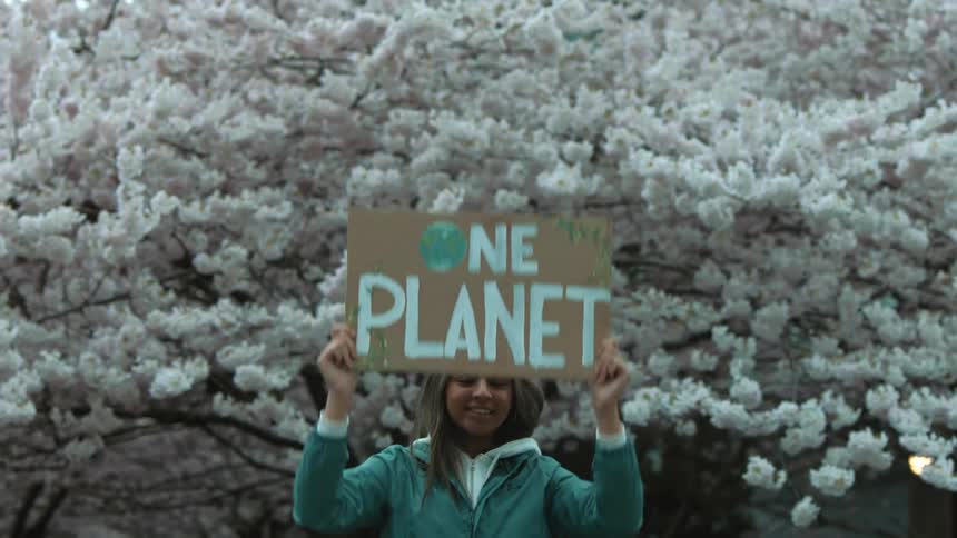 A young woman is holding up a climate change protest sign and standing by a tree