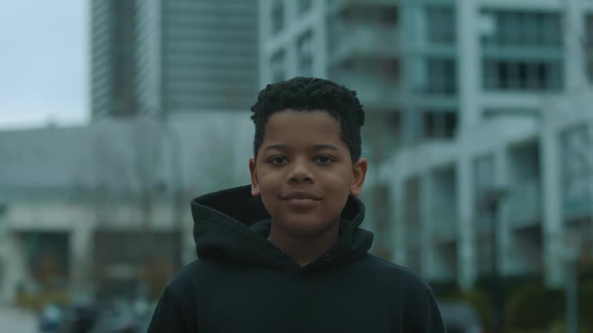 Young black boy with black hoodie smiles at camera in front of high rise building