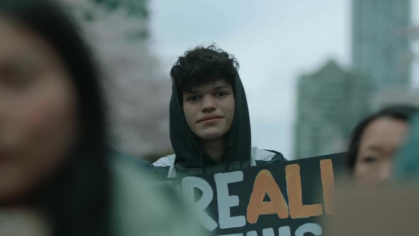 Young biracial man standing with a crowd holding a protest sign in the middle of a big city